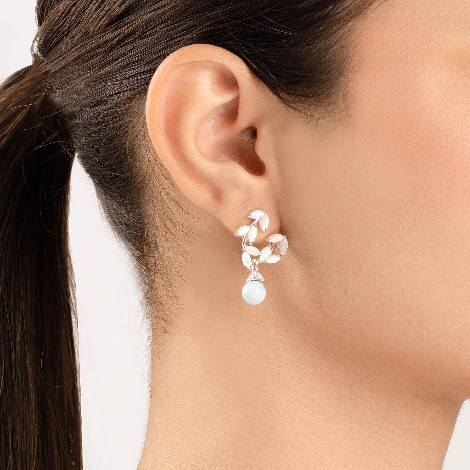 Close-up of an ear wearing a silver earring with a dangling pearl on a white background